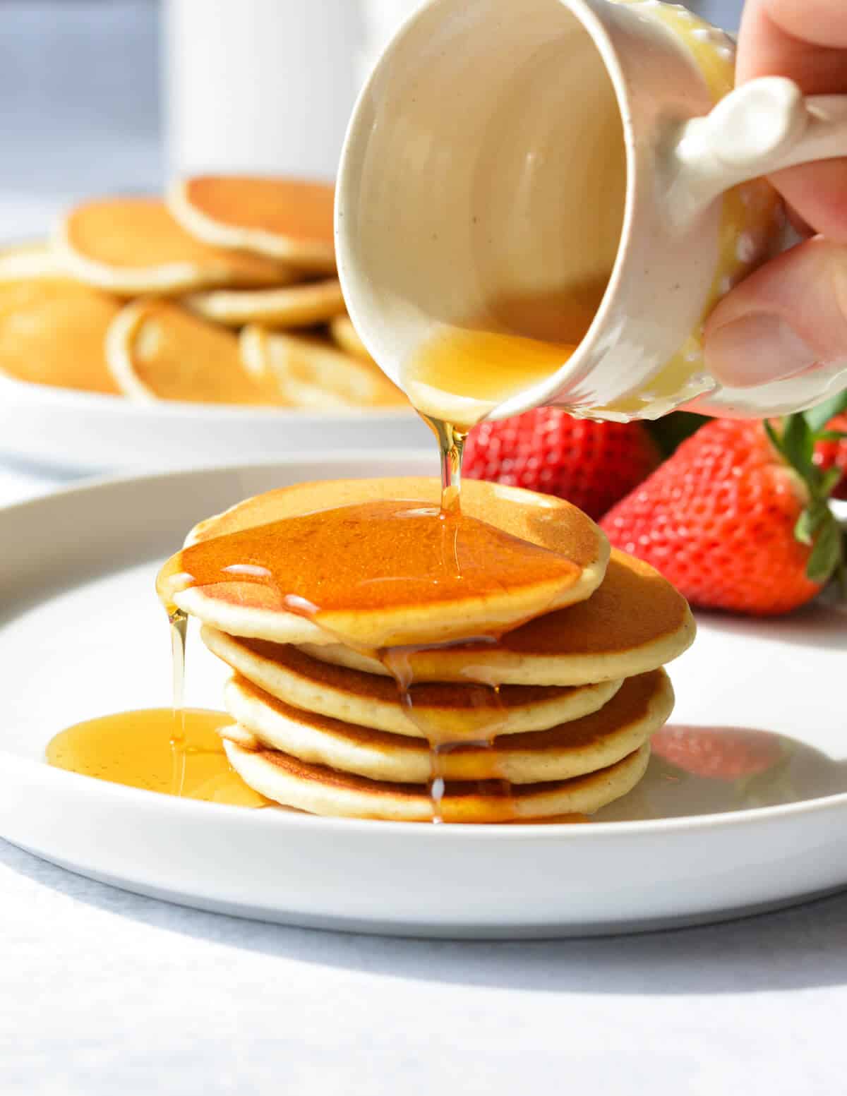 pouring maple syrup onto a stack of silver dollar pancakes. 